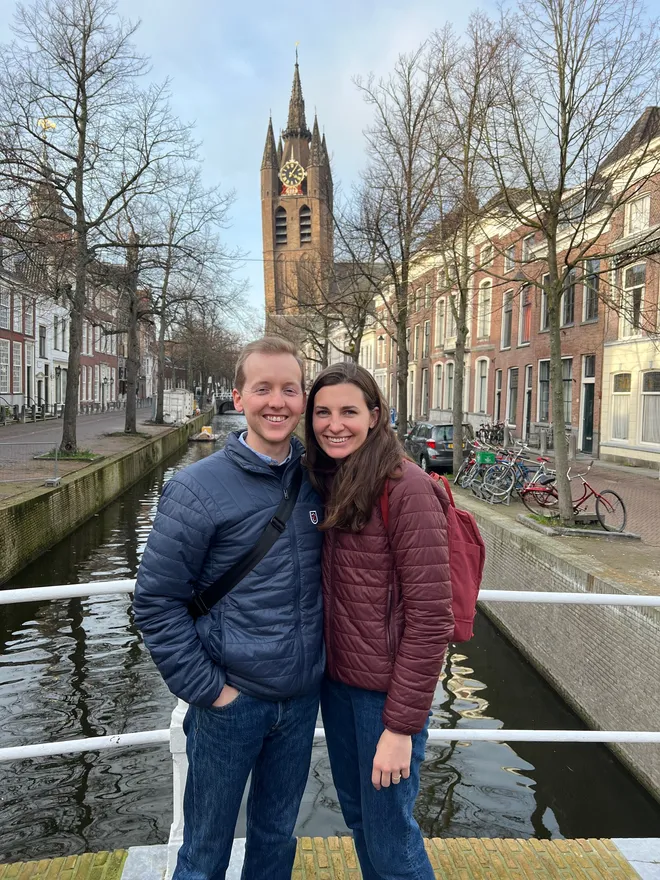 A couple smiling on a bridge over a canal, a brick church tower with a clock rising behind bare trees