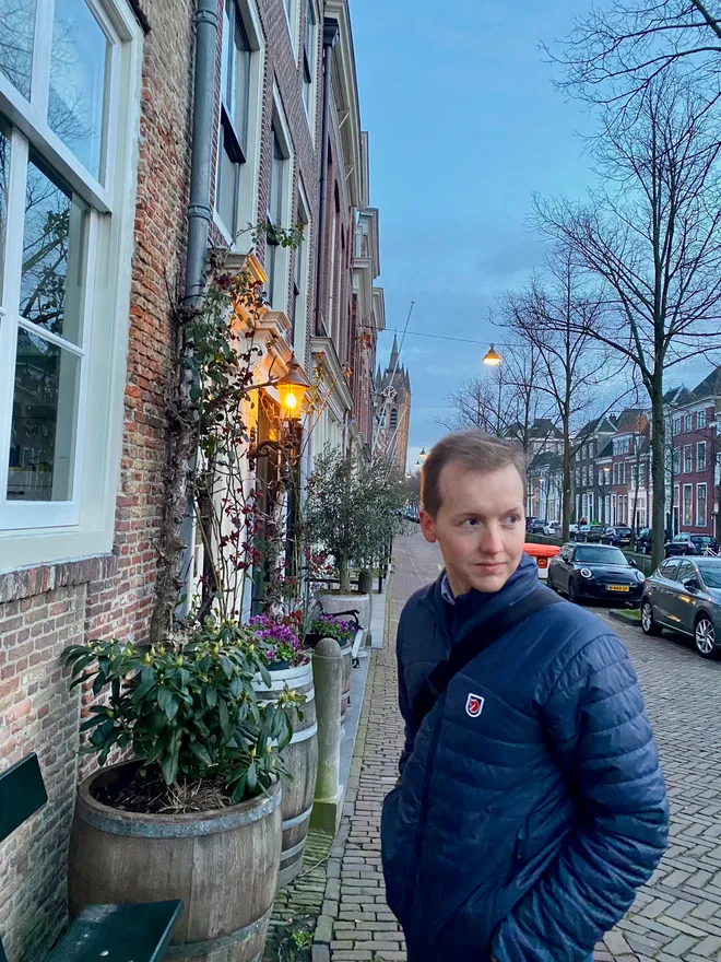A man in a navy puffer jacket glancing sideways on a brick street at dusk, a lit lantern and planters along the houses