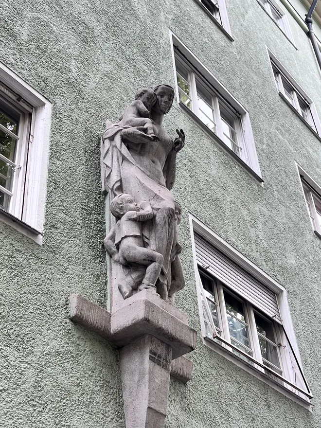 A stone sculpture of a woman with two children mounted on the corner of a green rendered apartment building