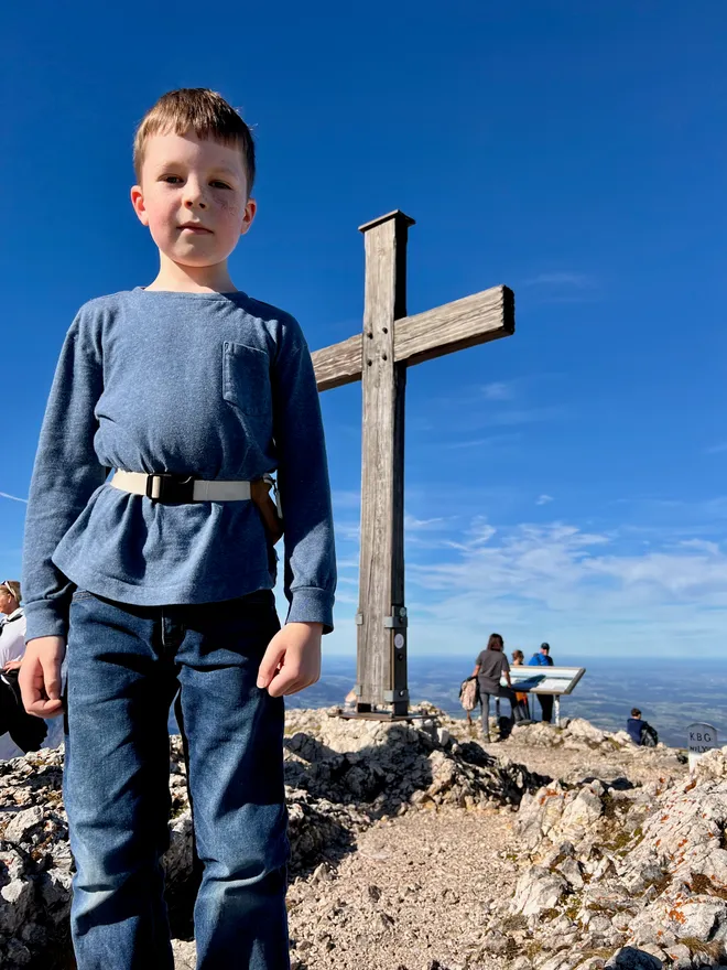 A boy photographed from below, standing beside a wooden cross against a deep blue sky