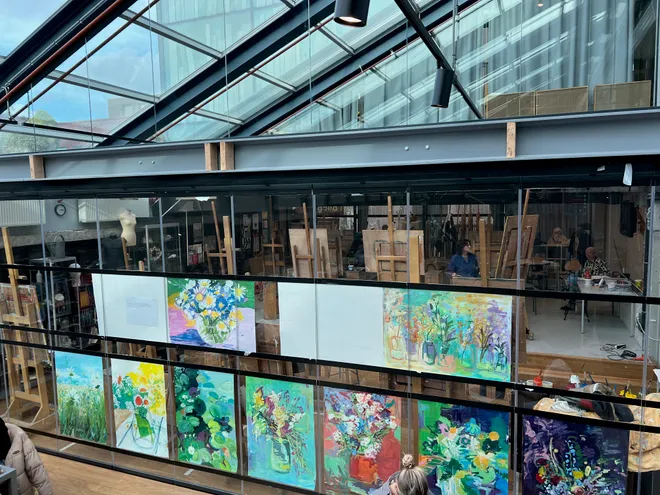 An open studio under a glass roof, canvases of flowers hung along a balcony rail above easels