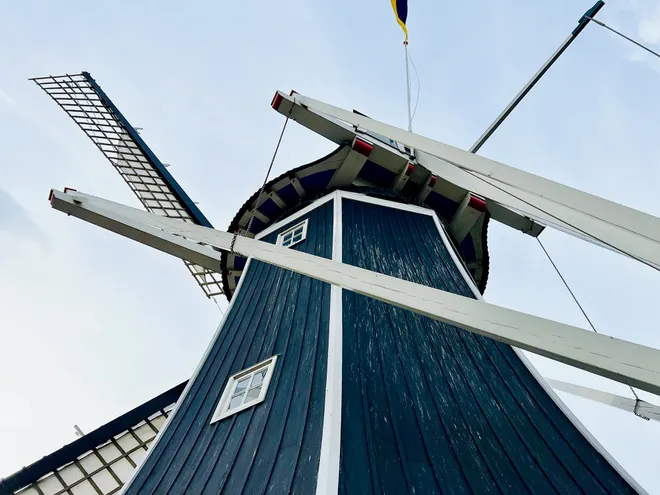 Looking up at a dark wooden windmill, its white sails crossing the frame against a pale sky